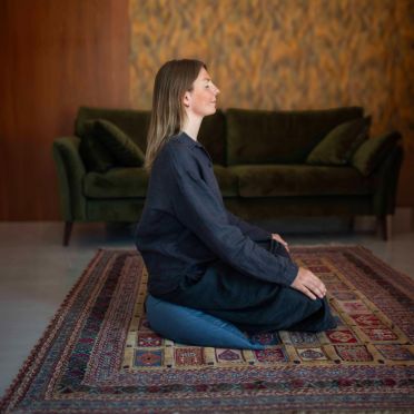 kobieta w ciemnych ubraniach siedzi na poduszce do medytacji i relaksacji w kształcie księżyca, w kolorze zgaszonym niebieskim. na podłodze leży wzorzysty dywan.